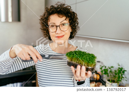 Smiling middle aged woman with curly hair and glasses cutting microgreens with scissors in kitchen, homegrown greens, eco lifestyle, plant based nutrition, healthy food preparation, indoor natural Smiling middle aged woman with curly hair and glasses cutting microgreens with scissors in kitchen, homegrown greens, eco lifestyle, plant based nutrition, healthy food preparation, indoor natural 138792017
