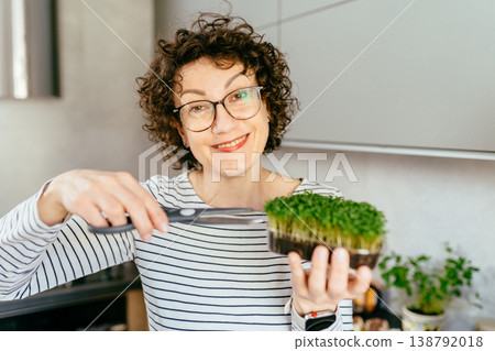 Smiling middle aged woman with curly hair cutting fresh microgreens, eco friendly lifestyle, plant based diet, home gardening, healthy food preparation in modern kitchen Smiling middle aged woman with curly hair cutting fresh microgreens, eco friendly lifestyle, plant based diet, home gardening, healthy food preparation in modern kitchen 138792018