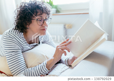 Close up female with glasses lying on bed and reading book. Focused calm mood, cozy indoor setting, quiet leisure, home reading routine, soft daylight, comfort and relaxation. 138792029