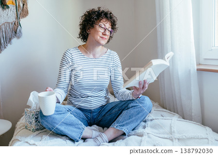 Female sitting on bed with book and mug, calm expression, quiet reading time, cozy bedroom interior, indoor rest, soft daylight, peaceful home routine and leisure moment. Female sitting on bed with book and mug, calm expression, quiet reading time, cozy bedroom interior, indoor rest, soft daylight, peaceful home routine and leisure moment. 138792030