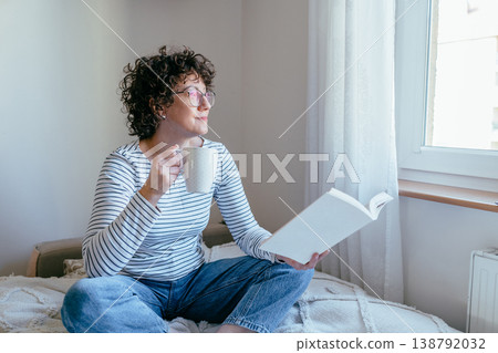 Curly haired female in her 40s with glasses holding cup and book, calm relaxed mood, daylight from window, indoor home environment, quiet personal routine Curly haired female in her 40s with glasses holding cup and book, calm relaxed mood, daylight from window, indoor home environment, quiet personal routine 138792032
