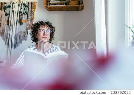 Curly haired female in her 40s with glasses holding book, thoughtful expression, indoor home setting, quiet reading moment, concentration and reflection 138792033