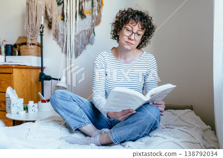Attractive confident curly haired female in her 40s with glasses reading book while sitting, relaxed calm expression, intellectual activity, indoor home environment, cozy routine Attractive confident curly haired female in her 40s with glasses reading book while sitting, relaxed calm expression, intellectual activity, indoor home environment, cozy routine 138792034