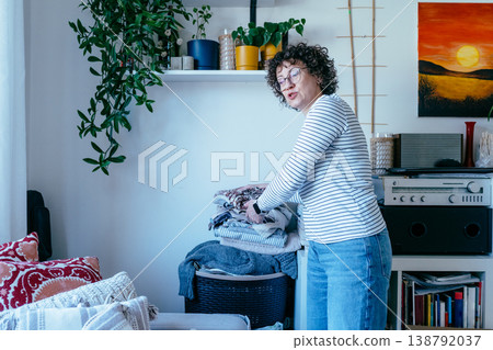 White woman in her 40s with curly hair placing folded clothes into basket, wardrobe organization, declutter process, home interior, clothing care, minimal lifestyle routine White woman in her 40s with curly hair placing folded clothes into basket, wardrobe organization, declutter process, home interior, clothing care, minimal lifestyle routine 138792037