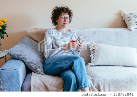 White woman in her 40s sitting on sofa using smartphone, relaxed slight smile, online communication, digital routine, home lifestyle, casual indoor moment, modern technology use White woman in her 40s sitting on sofa using smartphone, relaxed slight smile, online communication, digital routine, home lifestyle, casual indoor moment, modern technology use 138792041