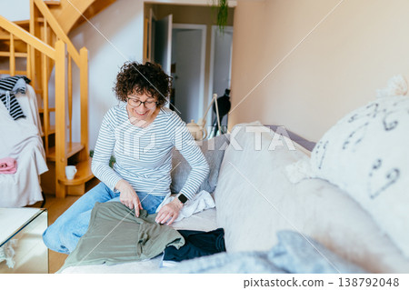 White woman in her 40s smiling while sorting green shirt on sofa, relaxed mood, wardrobe organization, home routine, clothing care, everyday domestic life, positive emotion White woman in her 40s smiling while sorting green shirt on sofa, relaxed mood, wardrobe organization, home routine, clothing care, everyday domestic life, positive emotion 138792048