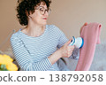 Middle aged woman with glasses gently removing lint from pink knitted sweater with fabric shaver. Calm focused mood, clothing care routine, cozy home environment, slow living concept. 138792058