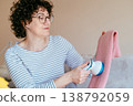 Middle aged woman with glasses holding pink sweater and using fabric shaver. Soft focus, relaxed mood, clothing care routine, gentle maintenance, cozy home lifestyle, everyday comfort. 138792059