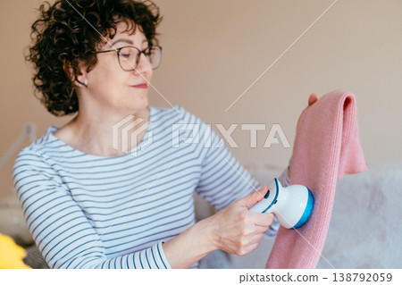 Middle aged woman with glasses holding pink sweater and using fabric shaver. Soft focus, relaxed mood, clothing care routine, gentle maintenance, cozy home lifestyle, everyday comfort. Middle aged woman with glasses holding pink sweater and using fabric shaver. Soft focus, relaxed mood, clothing care routine, gentle maintenance, cozy home lifestyle, everyday comfort. 138792059