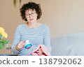 Middle aged woman with curly hair and glasses gently cleaning pink sweater with fabric shaver while sitting on sofa. Calm expression, home routine, clothing care, slow living, cozy domestic moment. 138792062