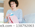 Middle aged woman with glasses carefully cleaning pink knitwear using fabric shaver. Concentrated expression, hands working on textile, clothing maintenance, pilling removal, everyday home routine. 138792063