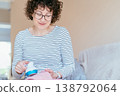 Middle aged woman with curly hair and glasses using fabric shaver on pink knitted sweater while sitting on sofa. Focused expression, clothing care, lint removal, home textile maintenance routine. 138792064