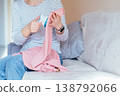 Unrecognizable woman cleaning pink knitted sweater with fabric shaver while sitting on sofa. Hands holding garment, removing pills, textile care, clothing maintenance, everyday domestic activity. 138792066