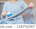 Unrecognizable woman in striped shirt stretching gray knitted blanket and removing lint with fabric shaver while sitting on sofa. Clothing care, pilling removal, textile maintenance at home routine. 138792067