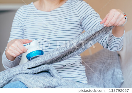 Unrecognizable woman in striped shirt stretching gray knitted blanket and removing lint with fabric shaver while sitting on sofa. Clothing care, pilling removal, textile maintenance at home routine. 138792067