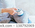 Close up female hand removing lint from gray knitted sweater with fabric shaver while sitting on sofa. Unrecognizable woman in striped shirt, home clothing care, pilling removal, textile routine. 138792069