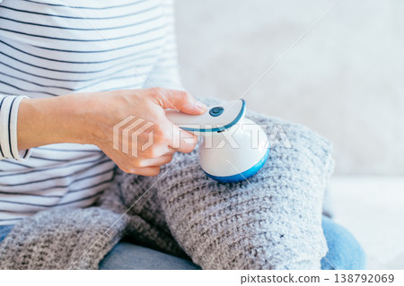 Close up female hand removing lint from gray knitted sweater with fabric shaver while sitting on sofa. Unrecognizable woman in striped shirt, home clothing care, pilling removal, textile routine. 138792069
