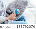 Cropped woman uses electric fabric shaver on gray knitted fabric while resting on sofa at home. Detail of hands, textile care, sweater cleaning, pilling removal, domestic routine, cozy maintenance. 138792070