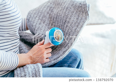 Cropped woman uses electric fabric shaver on gray knitted fabric while resting on sofa at home. Detail of hands, textile care, sweater cleaning, pilling removal, domestic routine, cozy maintenance. Cropped woman uses electric fabric shaver on gray knitted fabric while resting on sofa at home. Detail of hands, textile care, sweater cleaning, pilling removal, domestic routine, cozy maintenance. 138792070