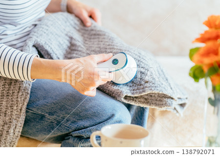 Close up of woman holding fabric shaver above gray knitted clothing during home care routine. unrecognizable person, soft textile, garment upkeep, lint removal, domestic maintenance. Close up of woman holding fabric shaver above gray knitted clothing during home care routine. unrecognizable person, soft textile, garment upkeep, lint removal, domestic maintenance. 138792071