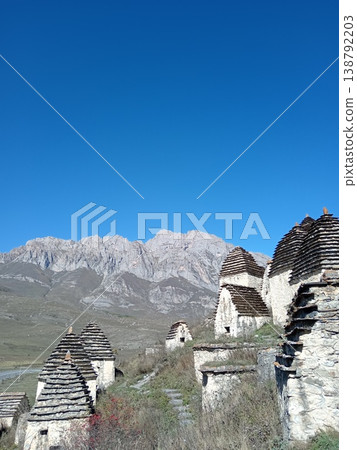 Dargavs necropolis, City of the Dead, North Ossetia, Russia. Rows of white stone tombs with pointed roofs against a backdrop of rugged Caucasus peaks 138792203