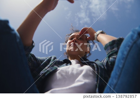 Low angle view of woman using smartphone outdoors against bright blue sky in sunny park environment Low angle view of woman using smartphone outdoors against bright blue sky in sunny park environment 138792857