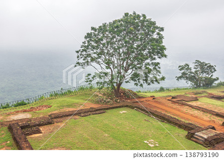Sigiriya ancient rock fortress in Sri Lanka. Ancient architectural ruins with green grass and lush trees atop a hill, overlooking a misty, verdant landscape Sigiriya ancient rock fortress in Sri Lanka. Ancient architectural ruins with green grass and lush trees atop a hill, overlooking a misty, verdant landscape 138793190