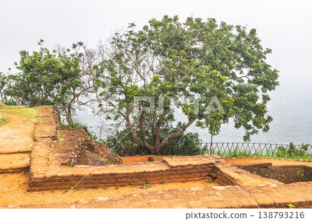 Sigiriya ancient rock fortress in Sri Lanka. Ancient Brick Foundations on Elevated Terrain with Lush Trees Against a Hazy Sky and Distant Hills Landscape 138793216