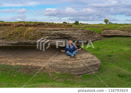Young woman sitting cross legged under stone ledge on grassy coast, calm posture, natural setting, confident body positive mood 138793924