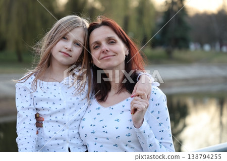A brunette mother with her 9-year-old blonde daughter in light-colored sweaters outside in the spring. Happy beautiful family. A brunette mother with her 9-year-old blonde daughter in light-colored sweaters outside in the spring. Happy beautiful family. 138794255