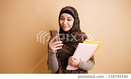 Muslim student smiling while checking smartphone and holding notebooks Muslim student smiling while checking smartphone and holding notebooks 138794340