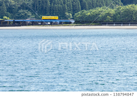 一列黃色的 Kiha 40 型列車駛過橫跨細田川的鐵路橋，細田川流入宮崎縣日南市的大土津海灘。 138794847