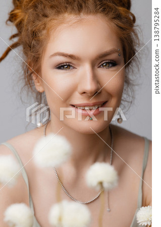 Young Caucasian redhead woman touching face among dried white flowers on moss, white wall 138795284