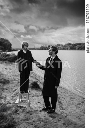 Father holding daughter hand standing on stool near river, cloudy sky, monochrome Father holding daughter hand standing on stool near river, cloudy sky, monochrome 138795309