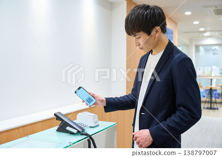 A young businessman scans a QR code on his smartphone at the office's reception system. A young businessman scans a QR code on his smartphone at the office's reception system. 138797007