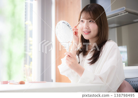 A close-up image of a woman applying blush to her cheeks while looking in a hand mirror, showcasing her natural makeup and beauty care routine. A close-up image of a woman applying blush to her cheeks while looking in a hand mirror, showcasing her natural makeup and beauty care routine. 138798495