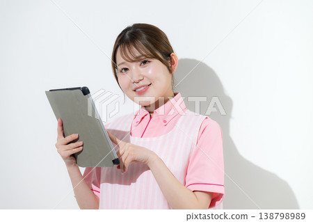 The image shows a woman working as a caregiver or childcare worker in a home care setting against a white background, pointing to a tablet as she engages in IT-related activities. The image shows a woman working as a caregiver or childcare worker in a home care setting against a white background, pointing to a tablet as she engages in IT-related activities. 138798989