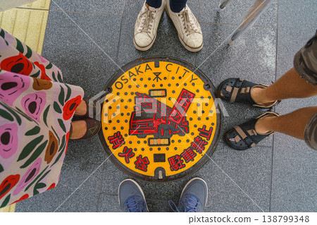 Osaka, Japan - Sep 22 2024, An overhead view of a manhole cover withred and yellow painting of fire truck, surrounded by the feet of four people, a man, a woman and a child, Osaka, Japan Osaka, Japan - Sep 22 2024, An overhead view of a manhole cover withred and yellow painting of fire truck, surrounded by the feet of four people, a man, a woman and a child, Osaka, Japan 138799348