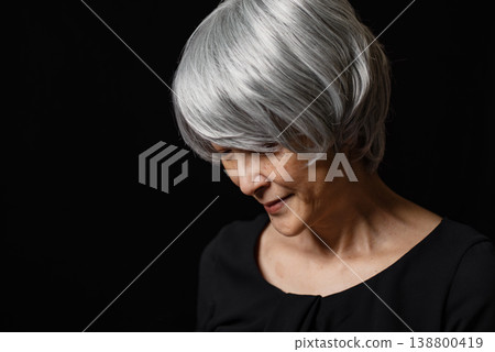 一位老婦人站在黑色背景前，低著頭，努力忍住笑。 138800419