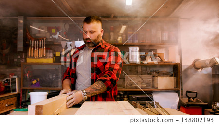 Joinery process. Stacking wood. Male carpenter checking plank quality holding timber on workbench in smoky workshop with equipment. 138802193