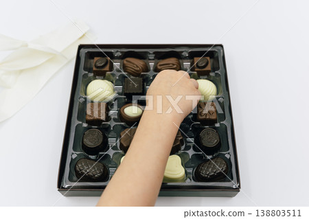 一隻孩子的手從放在白色背景的箱子裡拿起一塊巧克力。 一隻孩子的手從放在白色背景的箱子裡拿起一塊巧克力。 138803511
