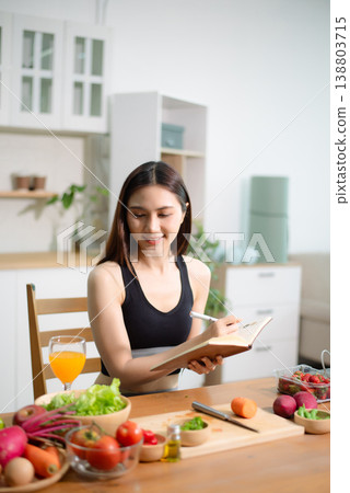 Asian woman reading recipe book while preparing fresh salad in home kitchen. Clean eating, nutrition planning, meal prep and healthy lifestyle concept. 138803715