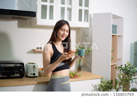 Asian woman in sportswear talking on smartphone while holding coffee at home kitchen. Healthy lifestyle, fitness routine, work life balance and modern living. Asian woman in sportswear talking on smartphone while holding coffee at home kitchen. Healthy lifestyle, fitness routine, work life balance and modern living. 138803769