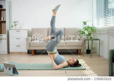Asian woman stretching on yoga mat at home while following online workout. Home yoga, fitness exercise, flexibility training, wellness lifestyle, indoor healthy living. 138803820