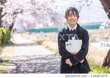 A girl in a school uniform stands on a road lined with cherry blossoms, holding a textbook. A girl in a school uniform stands on a road lined with cherry blossoms, holding a textbook. 138803979