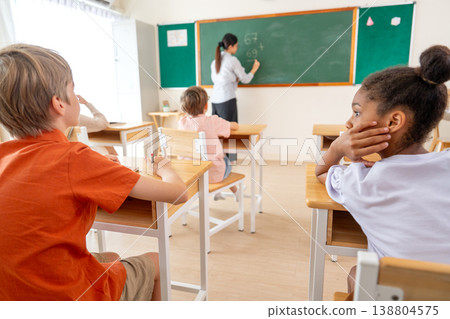 Vertical image of the teacher tell African American boy try to write the answer for calculation question on blackboard in front of classroom. 138804575