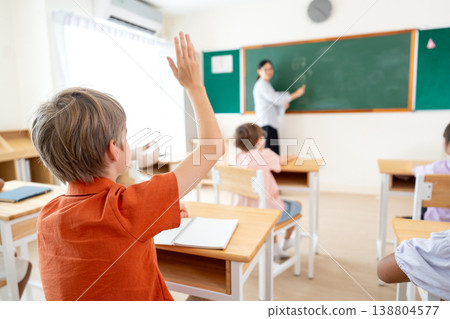 Back of the boy who raise his hand try to answer the calculation question that teacher write on board in classroom. Back of the boy who raise his hand try to answer the calculation question that teacher write on board in classroom. 138804577
