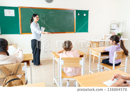 Asian teacher give colored chalk to little girl to write the answer of question on blackboard in front of classroom. Asian teacher give colored chalk to little girl to write the answer of question on blackboard in front of classroom. 138804578