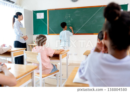 The teacher tell African American boy try to write the answer for calculation question on blackboard in front of classroom. The teacher tell African American boy try to write the answer for calculation question on blackboard in front of classroom. 138804580