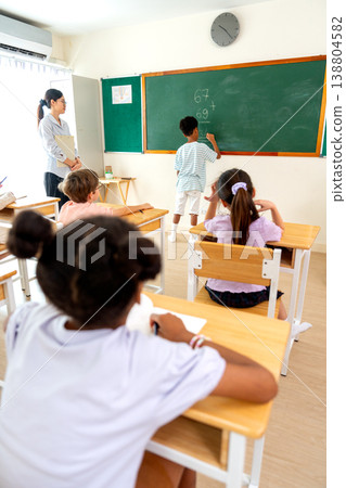 Vertical image of the teacher tell African American boy try to write the answer for calculation question on blackboard in front of classroom. Vertical image of the teacher tell African American boy try to write the answer for calculation question on blackboard in front of classroom. 138804582